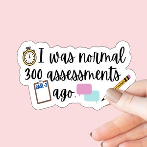 May include: A white sticker with black text that reads "I was normal 300 assessments ago." The sticker features a small clock, a clipboard with the text "CASL-2", a pink speech bubble, and a blue speech bubble. A pencil is shown in the background.