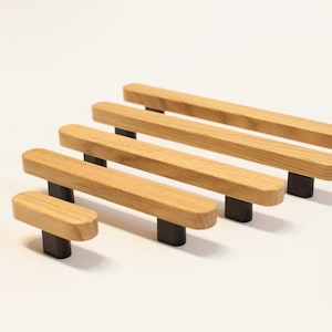 以下が含まれることがあります： さまざまなサイズの木製キャビネットプル。各プルは、濃い灰色の長方形のベースに取り付けられた、明るい茶色の長方形の木製ハンドルを備えています。白い表面に配置されています。