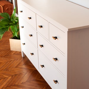 Peut inclure: Une commode blanche avec neuf tiroirs et des poignées de tiroir noires et en bois. La commode est dans une pièce avec un parquet.