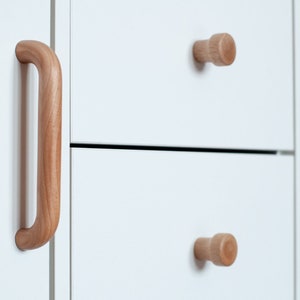 Könnte beinhalten: Ein weißer Schrank mit zwei Schubladen und einer Tür. Die Schubladen haben Holzknöpfe und die Tür einen langen Holzgriff. Der Schrank steht an einer Wand mit einem Bücherregal rechts.