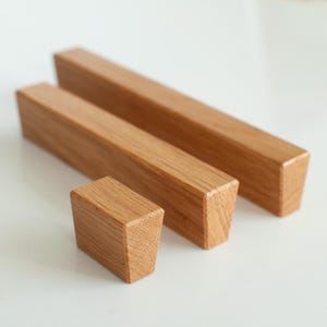 Könnte beinhalten: Drei Holzblöcke unterschiedlicher Größe, angeordnet auf einer weißen Oberfläche. Die Blöcke sind rechteckig mit natürlicher Holzmaserung. Der kleinste Block ist ein Würfel, während die anderen längere, rechteckige Formen haben.