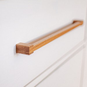 Peut inclure: Gros plan sur une poignée de tiroir en bois de forme rectangulaire. La poignée est en bois brun clair et est fixée à un tiroir blanc.