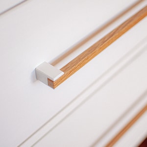 Op de afbeelding: Een witte lade met een houten handvat. Het handvat is bevestigd aan de lade met een witte metalen beugel.