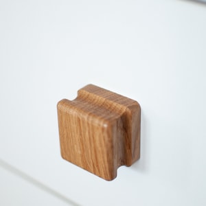 Può includere: Una maniglia per cassetto quadrata in legno con una sezione centrale rialzata. La maniglia è in legno marrone chiaro con una venatura naturale.