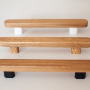 Könnte beinhalten: Drei Holzschrankgriffe mit natürlicher Holzmaserung. Ein Griff hat weiße Stützen, einer Holzstützen und einer schwarze Stützen. Die Griffe sind rechteckig mit abgerundeten Kanten.