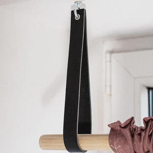 May include: A black leather strap suspended from a silver metal hook, looped around a light-coloured wooden rod. The rod holds a gathered, dark mauve fabric curtain. The strap is secured at the top with a silver screw and metal hardware.