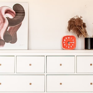 May include: A white dresser with six drawers and gold-coloured knobs. The dresser has a framed abstract print above it and a red clock and a vase with dried flowers on top.