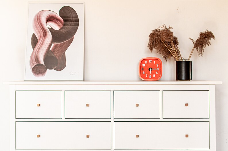 May include: A white dresser with six drawers and gold-coloured knobs. The dresser has a framed abstract print above it and a red clock and a vase with dried flowers on top.