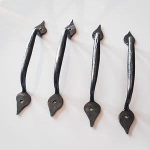 Peut inclure: Quatre poignées de porte en fer forgé noir, avec une base en forme de goutte d'eau et un fleuron décoratif. Les poignées sont incurvées et présentent un aspect texturé et martelé, sur fond blanc.