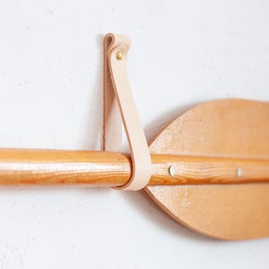 Op de afbeelding: Een houten kanopeddel met een lichte houtnerf hangt horizontaal aan een enkele, bleekbeige lederen riem. De riem is vastgezet met een kleine, messingkleurige metalen klinknagel, waardoor de peddel stevig tegen het muuroppervlak wordt gehouden.