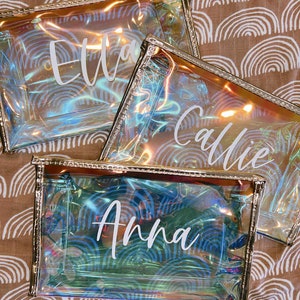 K&ouml;nnte beinhalten: Drei durchsichtige, schimmernde Kosmetiktaschen mit goldfarbenem Besatz. Die Taschen sind mit den Namen "Ella", "Callie" und "Anna" in wei&szlig;er Schrift personalisiert.