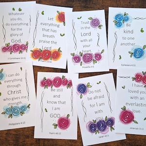 May include: A collection of white cards with inspirational quotes and floral illustrations. Each card features a different quote in black text, accompanied by watercolor-style roses in various colors. The cards are arranged on a wooden surface.