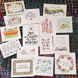 May include: A collection of fourteen rectangular greeting cards featuring hand-lettered inspirational quotes and colorful illustrations including floral wreaths, bees, hearts, and rainbows. Each card displays unique religious text and vibrant watercolor accents.