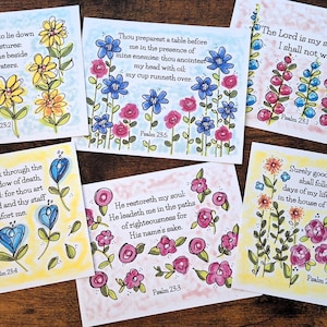 May include: A collection of six watercolor art prints featuring floral illustrations and Bible verses. Each print showcases colorful flowers in various arrangements, with text from Psalms. The prints are on a wooden surface.