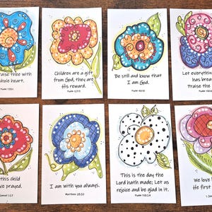 May include: Set of eight floral art cards with colorful, hand-drawn flower illustrations. Each card features a unique flower design and a religious quote. The cards are on a wooden surface.