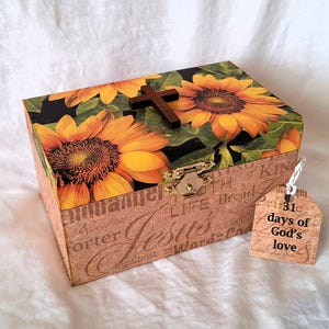 May include: A wooden box with a sunflower design and a cross on the lid. The box is decorated with a brown paper background with words and phrases in white. A tag attached to the box reads "31 days of God's love".