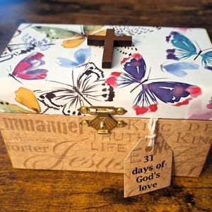 May include: A decorative wooden box with a butterfly design on the lid and a small wooden cross. The box has a gold clasp and a tag that reads "31 days of God's love". The box is on a wooden surface.
