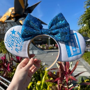 May include: Blue sequined bow headband with white ear details. The ears feature the text "Nobody's Gonna Stop Rock n' Roll From Exi" and a cassette tape design. The headband is held up against a sunny outdoor background.
