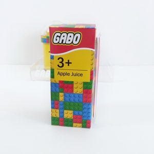 May include: A yellow cardboard box with a red and yellow logo that says "Gabo" and "3+ Apple Juice". The box is decorated with a pattern of colorful Lego bricks.