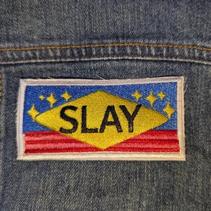 Peut inclure: Écusson brodé avec le mot "SLAY" en lettres majuscules noires. L'écusson présente une forme de losange jaune, un fond bleu avec des étoiles jaunes et des rayures rouges et blanches. L'écusson est cousu sur une veste en jean.