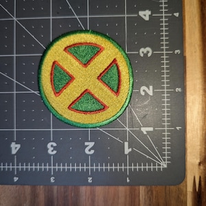 May include: Embroidered patch with a yellow circular base and a green and red X-shaped design. The patch has a stitched border. The patch is on a black cutting mat with metric and imperial markings.