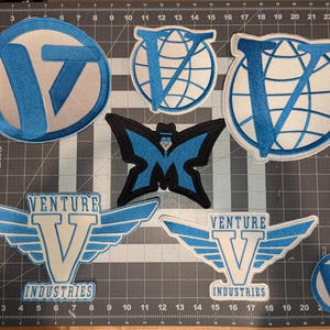 May include: Six embroidered patches featuring a blue "V" with white accents. The patches include a butterfly, a globe, and two versions of a logo that says "Venture Industries".