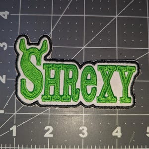 May include: Embroidered patch featuring the word "SHREXY" in green, with a black outline, on a white base. The design has a distinctive "S" and a fun font. This patch is suitable for application on clothing or accessories.