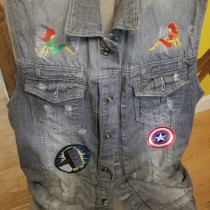 May include: A gray denim vest with embroidered patches. The patches include a red and yellow woman, a green and yellow woman, a blue and yellow hammer, and a red, white, and blue star.