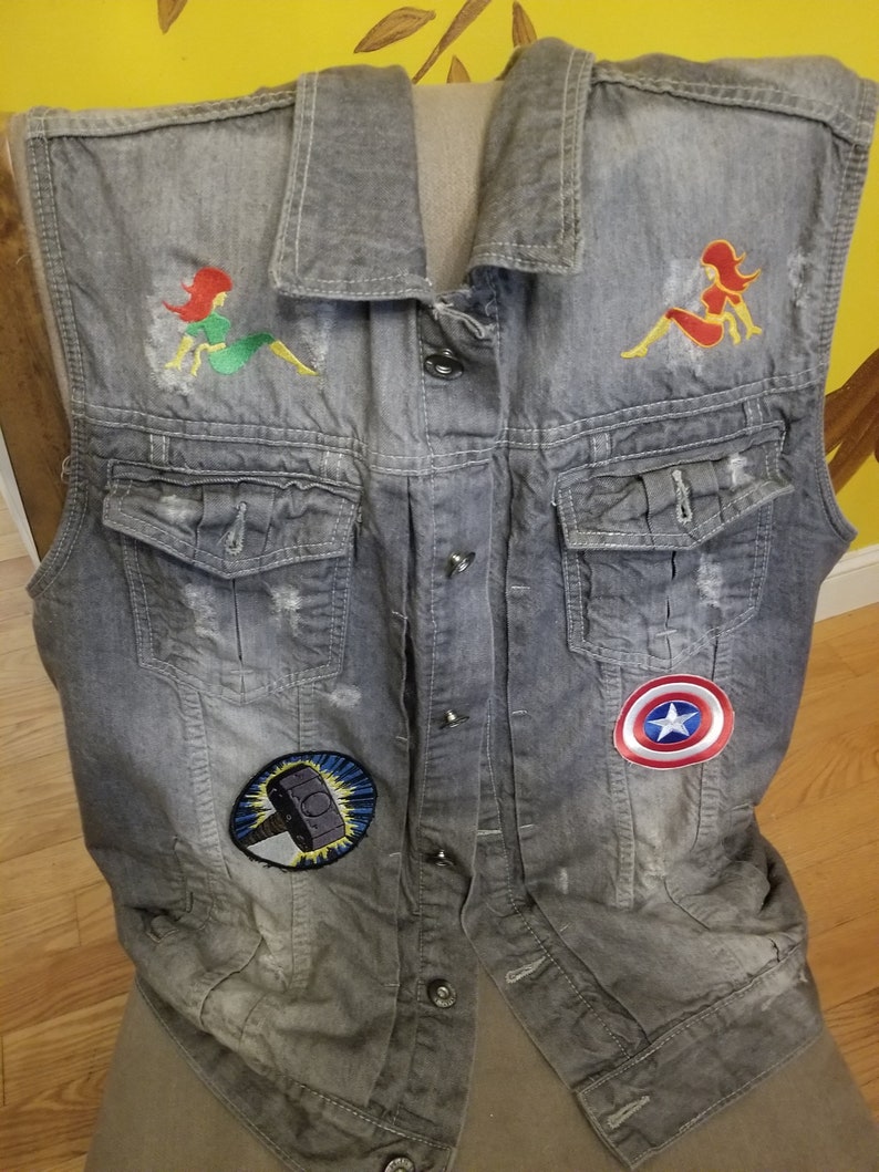 May include: A gray denim vest with embroidered patches. The patches include a red and yellow woman, a green and yellow woman, a blue and yellow hammer, and a red, white, and blue star.