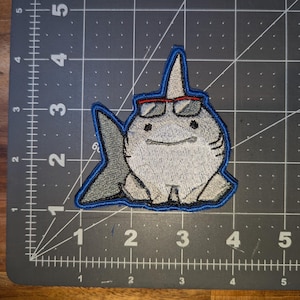 May include: Embroidered patch featuring a cartoon shark wearing sunglasses. The shark is gray with a white belly, a blue outline, and a fin. The sunglasses have red frames. The patch is approximately 3 inches wide.