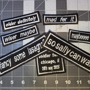 May include: A collection of black and white embroidered patches with various phrases, including "older definitely," "wiser maybe," and "so sally can wait." The patches are rectangular with white borders and text.