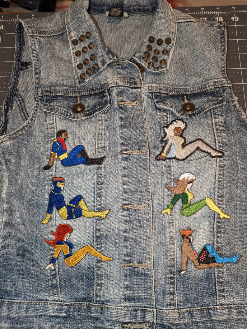 May include: A denim vest with a studded collar and embroidered patches of various characters from the X-Men comic book series. The patches are in a variety of colors, including blue, yellow, red, green, and black.