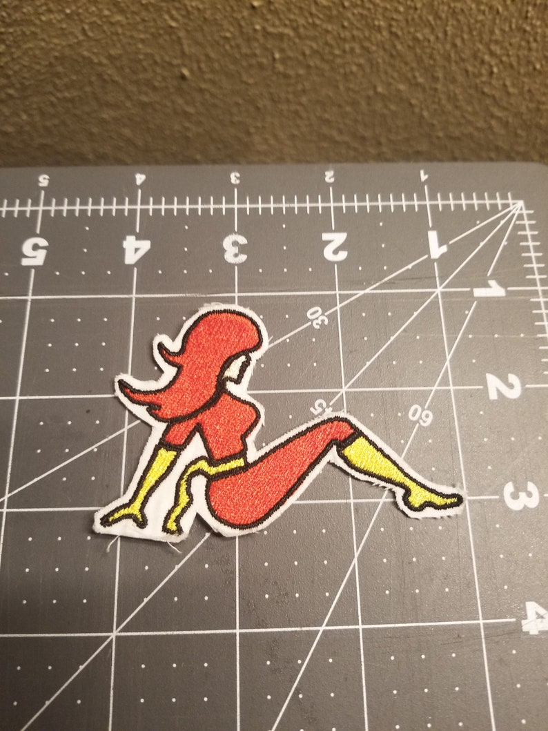 May include: Red and yellow embroidered iron-on patch of a woman sitting with her legs crossed.