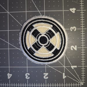 May include: Circular embroidered patch with a black and cream geometric design. The patch has a black border and a cream-colored center with a black cross-like pattern. The patch is on a gray cutting mat.