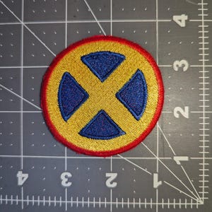 May include: Embroidered patch featuring a yellow circle with a blue X design, outlined in red. The patch is on a gray cutting mat with a grid pattern. This patch is suitable for jackets, bags, or other fabric items.