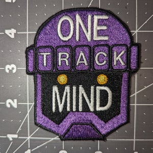 Peut inclure: Patch brod&eacute; violet avec une t&ecirc;te de robot et le texte "ONE TRACK MIND" en blanc.