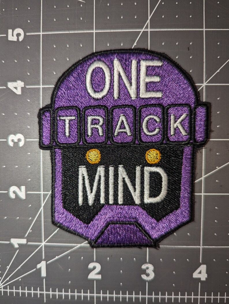 Peut inclure: Patch brod&eacute; violet avec une t&ecirc;te de robot et le texte "ONE TRACK MIND" en blanc.