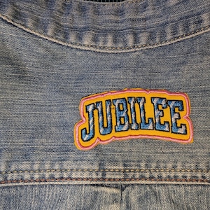 May include: A light blue denim jacket featuring an embroidered patch on the back. The patch displays the word JUBILEE in blue letters with a yellow background and a pink border, stitched directly onto the fabric of the denim garment.