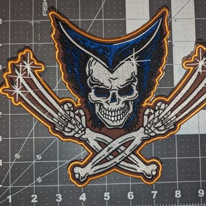 May include: A brown and yellow embroidered patch featuring a skull with blue and black hair, crossed bone claws, and a white background.
