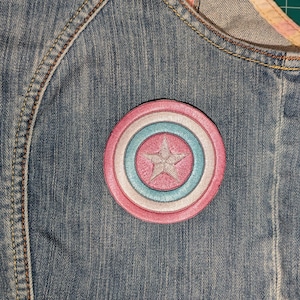 May include: A round, embroidered patch with a star design, featuring pink, light blue, and white concentric circles. The patch is affixed to a denim garment, showcasing a classic superhero theme.