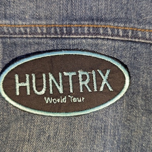 May include: An embroidered patch with the word "HUNTRIX" in light blue lettering on a black oval. The text "World Tour" is below. The patch is sewn onto a blue denim jacket.