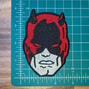 May include: Embroidered patch featuring a stylized face with red, black, and off-white colors. The design includes a mask, horns, and a determined expression. The patch is placed on a green cutting mat with measurement markings.