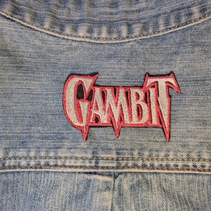 May include: A light blue denim jacket featuring an embroidered patch on the back. The patch displays the word GAMBIT in a stylized silver font with a bold red outline, stitched directly onto the fabric with visible horizontal denim texture.