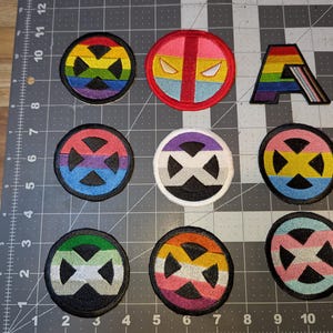 May include: A collection of nine colorful embroidered patches. The patches feature various designs, including a rainbow letter "A", and circular emblems with geometric shapes and rainbow, pink, blue, and yellow color schemes. The patches are on a gray cutting mat.