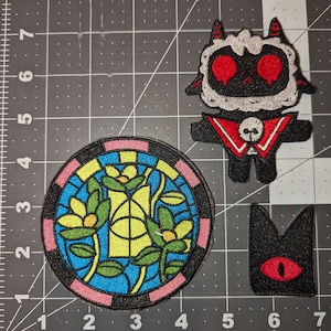 May include: Three embroidered iron-on patches. A black and white sheep with red eyes and a red collar with a skull and crossbones. A round stained glass window with yellow flowers and green leaves. A black cat with a red eye.