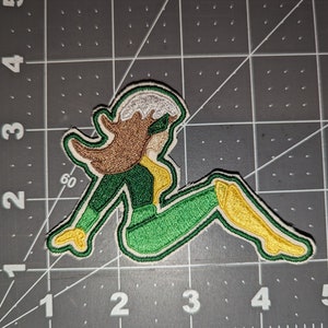 May include: Green and yellow embroidered patch of a cartoon superhero sitting with their legs crossed. The superhero has brown hair and a white headband.