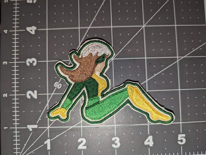 May include: Green and yellow embroidered patch of a cartoon superhero sitting with their legs crossed. The superhero has brown hair and a white headband.