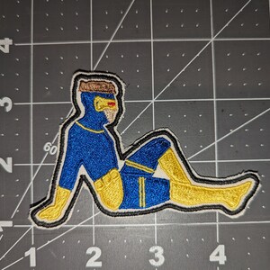 May include: A blue and yellow embroidered patch of a person in a superhero pose. The person is wearing a blue suit with yellow accents and a yellow mask.