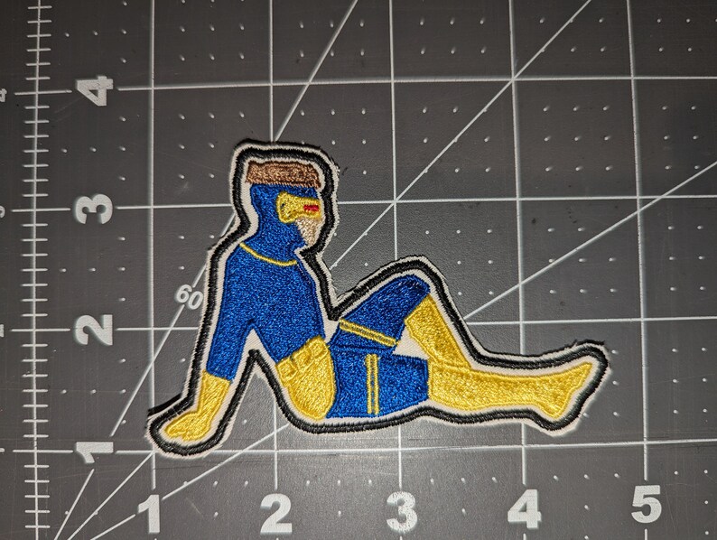 May include: A blue and yellow embroidered patch of a person in a superhero pose. The person is wearing a blue suit with yellow accents and a yellow mask.