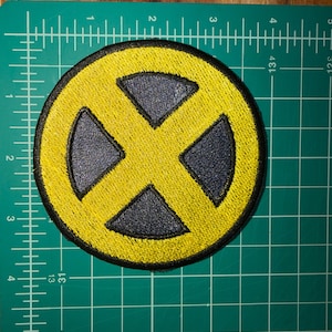 May include: A circular embroidered patch featuring a yellow X symbol centered on a dark blue background, enclosed by a thick black border. The patch is positioned on a green cutting mat with white grid lines, showing a diameter of 3 inches.